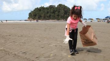 criança usa luva para pegar um microlixo na faixa de areia. Ela segura um saco de papel para depositar o resíduo.#paratodosverem