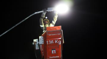 #pracegover Erguido dentro de cabine, homem de uniforme e luvas manuseia lâmpada de poste público à noite