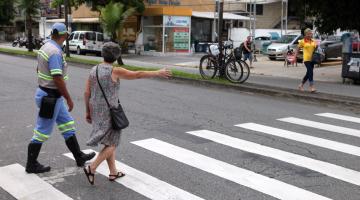 Mulher faz o sinal típico da Faixa Viva. Ela está percorrendo uma faixa de pedestres. Agente da CET está ao lado dela. #Pracegover