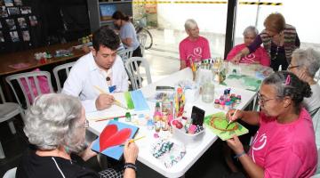 idosos estão sentados em torno de uma mesa pintando telas. A mesa está cheia de apetrechos como tintas e pinceis. #paratodosverem 