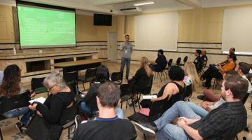 pessoas estão sentadas em auditório assistindo apresentação. Um homem fala a todos. #paratodosverem