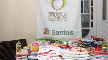 pacotes de alimentos estão sobre uma mesa. Há um banner do fundo social ao fundo. #paratodosverem 