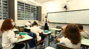 alunos sentados às carteiras estão usando máscara. Ao fundo, a professora à frente do quadro. #paratodosverem