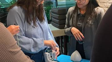 mulher está diante de uma mesa mexendo e apresentando dispositivo urinário para outras pessoas.  #paratodosverem 