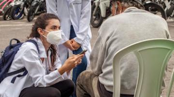 Profissionais de saúde, uma em pé e outra agachada, conversam com idoso sentado em cadeira. #pracegover