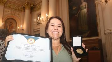 mulher segura diploma com a mão direita e medalha com a esquerda. #paratodosverem