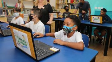 crianças estão sentadas em mesas diante de laptops. Em primeiro plano, o computador está aberto. Na tampa  se vê o nome do programa Grêmio Estudantil. #paratodosverem