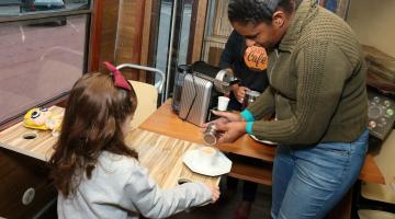 Mulher está em pé adiciona pó (canela ou chocolate) em xícara de café. Uma  menina, de costas para a imagem e com laço na cabeça, está segurando outra xícara de café. #Pracegover