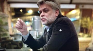 O ator fábio assunção, personagem da série, está sentado num balcão de bar. Ele está em primeiro plano. O fundo está desfocado. #paratodosverem