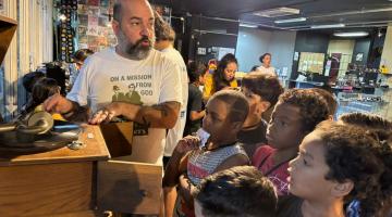 homem apresenta vitrola antiga de madeira para crianças reunidas em torno dele. #paratodosverem