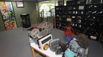 área interna de museu. com mesa e vários objetos antigos, como câmeras e filmadoras sobre uma mesa. Ao fundo, prateleiras com objetos de fotografia. #paratodosverem