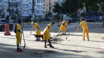 Operários uniformizados varrem trecho de pista . A área onde fazem o serviço está demarcada por cones. Eles usam vassoura e um carrinho de  mão. Usam máscaras e luvas. #Paratodosverem