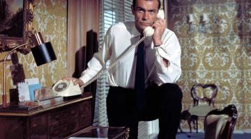 James Bonde está ao telefone em um quarto. Uma de suas pernas está apoiada sobre uma cadeira. #Pracegover