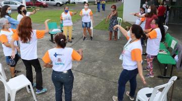 ATIVIDADE AO AR LIVRE. VÁRIAS PESSOAS ESTÃO EM PÉ, EM RODA, FAZENDO MOVIMENTOS COM OS BRAÇOS. #PARATODOSVEREM