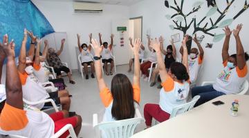 idosos estão sentados em cadeiras reunidos em roda. Todos fazem movimentos com os braços para o alto. #paratodosverem