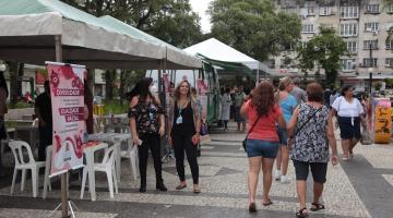 tendas armadas na praça à esquerda. Mulheres em frente às tendas e circulando. #paratodosverem