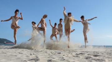 Bailarinas saltam na areia em dia ensolarado sem nuvens no céu azul ao fundo. #pracegover