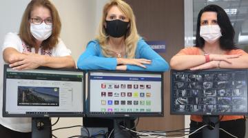 Três mulheres usando máscaras estão em pé com os braços cruzados e pousados sobre os respectivos desktopos - #Paratodosverem