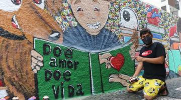 Mural grafitado com livro aberto onde se lê Doe Amor doe vida. O artista etá agachado no chão, segurando uma lata de tinta spray. #paratodosverem