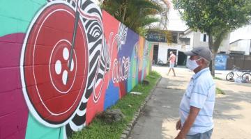 homem em pé na praça usando máscara olha para pintura em muro. . Trecho da calçadaé ajardinado. #paratodosverem