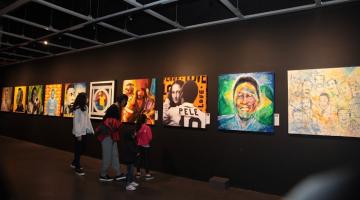 Interior do Museu Pelé com fotos e pinturas do Rei feitas por artistas. Há público observando. #paratodosverem