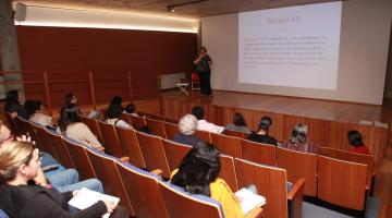 Mulher dá palestra sobre palco de auditório do Museu Pelé, assistido por público sentado. #pracegover