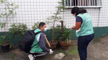 dois agentes estão no quintal de uma moradia. O rapaz está agachado próxima a alguns vasos. A moça está em pé fazendo anotação em uma prancheta. #paratodosverem