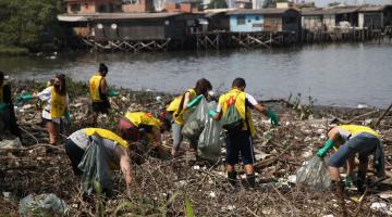 pessoas uniformizadas estão em área de mangue, próximo à água, limpando o terreno. #paratodosverem