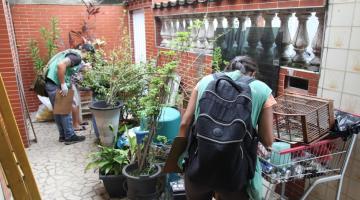 Agentes estão na parte externa da frente de uma casa onde procuram focos de larvas. A área tem muitas plantas e quinquilharias. #paratodosverem