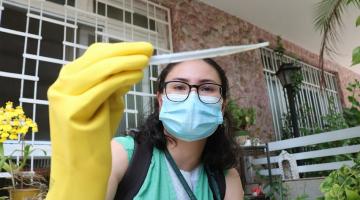 agente de saúde uniformizada e usando luva e máscara segura frasco com larvas recolhidas. Atrás se vê a frente de uma casa com plantas. #paratodosverem