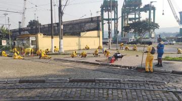 vários homens vestindo uniformes amarelos fazem capinação em calçada. Ao fundo, guindastes no porto. #paratodosverem