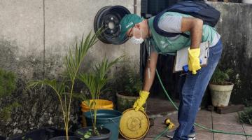 agente uniformizado se curva para pegar prato de planta junto a um jardim. Ele usa luvas nas duas mãos. #paratodosverem