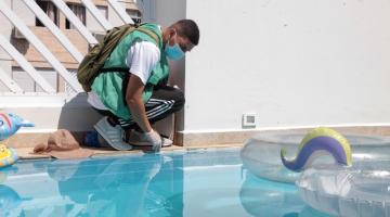 agente uniformizado se ajoelha ao lado de piscina e aproxima frasco da linha d'água. Há boias ao lado direito da piscina. #paratodosverem