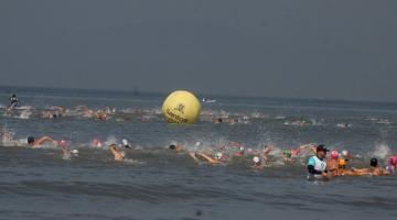 atletas estão nadando no mar. Ao fundo, uma grande boia de marcação onde se vê o brasão e o nome da cidade. #paratodosverem 