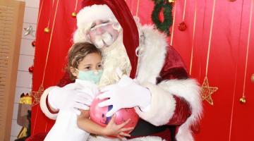 Papai Noel abraça menina. Os dois seguram uma bola colorida. #paratodosverem