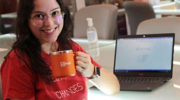 jovem sorri para a foto segurando uma caneca. Ela está sentada e há um computador pessoal atrás dela. #paratodosverem