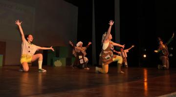 artistas apresentam peça infantil em palco. Eles estão com um dos joelhos apoiados no chão e um dos pés. As mãos estão para o alto. Todos sorriem. #paratodosverem