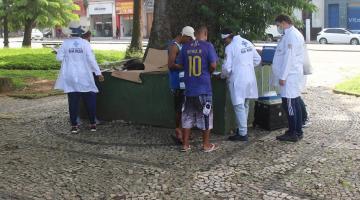 agentes usando avental branco prestam atendimento a uma pessoa em praça pública. #paratodosverem