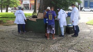 agentes usando avental branco prestam atendimento a uma pessoa em praça pública. #paratodosverem