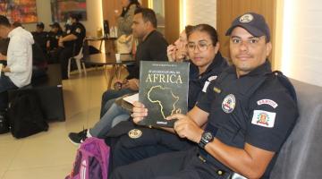 guarda municipal está sentado segurando livro em cuja capa se lê Africa e se vê o desenho do continente africano. #paratodosverem