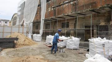 homem está trabalhando em canteiro de obras à frente de uma grande fachada com andaimes. #paratodosverem