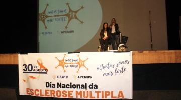 faixa à frente de palco onde se lê Dia Nacional da Esclerose Múltipla. No palco há duas mulheres, uma delas em cadeira de rodas. #paratodosverem