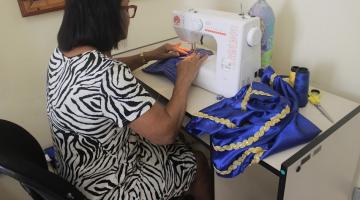 mulher está de costas para a foto confeccionando peça em máquina de costura. Ao lado, um tecido de cetin em azul e dourado. #paratodosverem