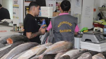 fiscal está de costas conversando com homem em box de peixe. à frente, um balcão cheio de peixes. #paratodosverem 