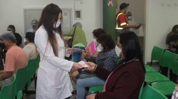 mulher usando avental branco entrega folheto para outra mulher que está sentada em sala de espera. Outras pessoas estão sentadas também. #paratodosverem