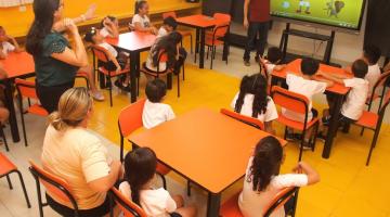 professor está diante de tela apontando para figura e crianças estão sentadas prestando atenção. #paratodosverem