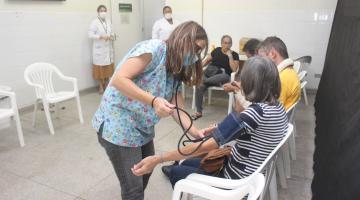 mulher mede a pressão de outra mulher que está em pé. Há outras pessoas sentadas na sala. #paratodosverem