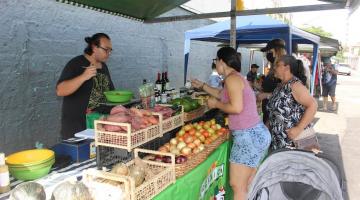 comerciante está à frente de banca de legumes. Consumidores estão olhando e tocando. #paratodosverem 