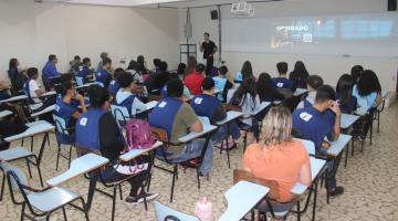 pessoas estão sentadas em sala de aula. todos estão de costas para a foto. Ao fundo, um homem fala ao grupo à frente de um telão. #paratodosverem