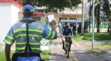 agente de trânsito está de costas segurando uma plaquinha. Ele está ao lado da ciclovia, por onde passam ciclistas. #paratodosverem 